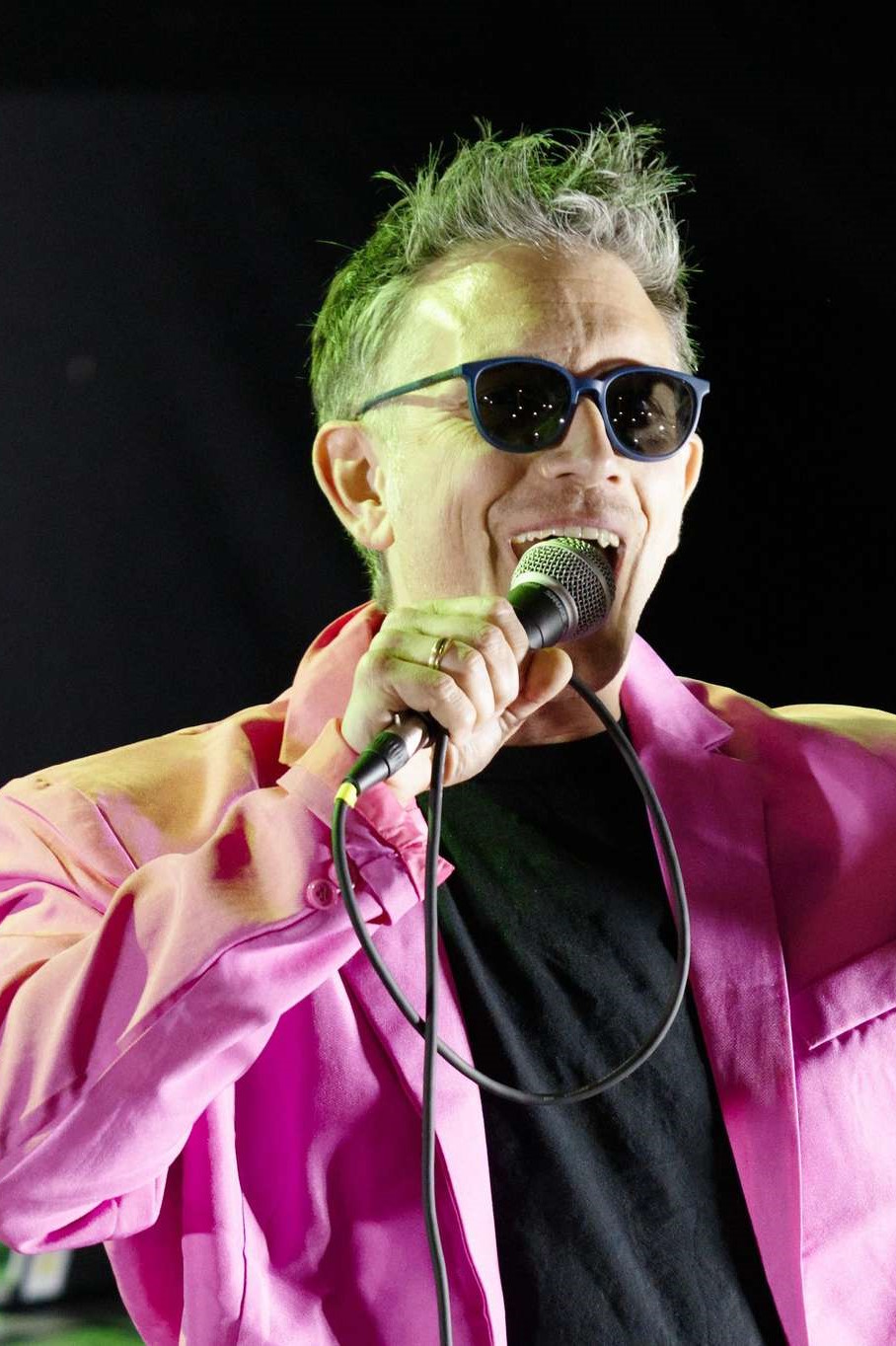 Mann mit grauem Haar, dunkler Sonnenbrille und pinkfarbenem Sakko singt leidenschaftlich in ein Mikrofon vor schwarzem Hintergrund, grünes Bühnenlicht fällt von oben. In den Brillengläsern spiegeln sich Handylampen. (Foto von Don Clemente)